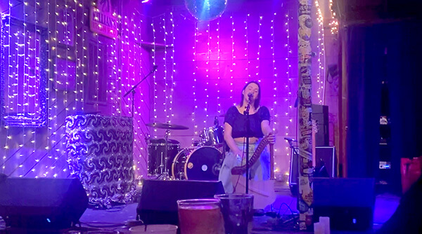 Musician sitting and playing a harp on a stage decorated with string lights and drums in the background.