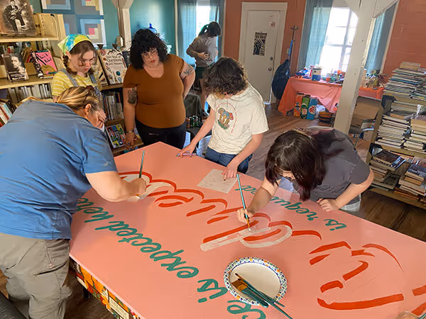 Five people painting a large pink sign with red and green letters in an indoor room filled with books and posters.