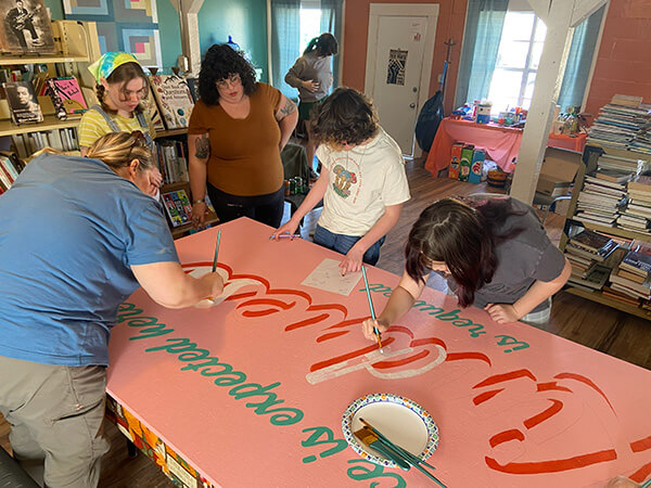Five people painting a large pink sign with red and green letters in an indoor room filled with books and posters.