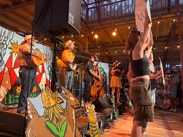 Band performing on a decorated wooden stage with painted animal and floral cutouts, while a crowd watches inside a wooden-beamed venue.