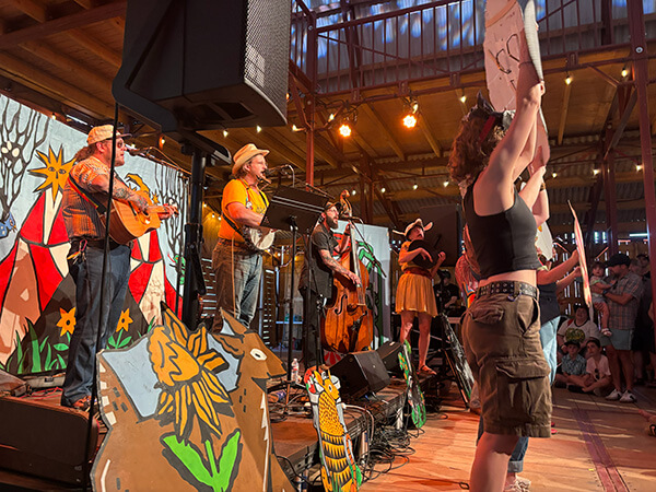 Band performing on a decorated wooden stage with painted animal and floral cutouts, while a crowd watches inside a wooden-beamed venue.