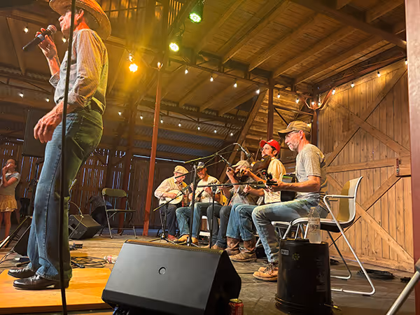 String band performing on stage in a wooden barn with musicians playing instruments and a singer wearing a cowboy hat.