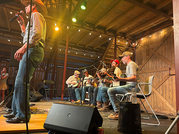 String band performing on stage in a wooden barn with musicians playing instruments and a singer wearing a cowboy hat.