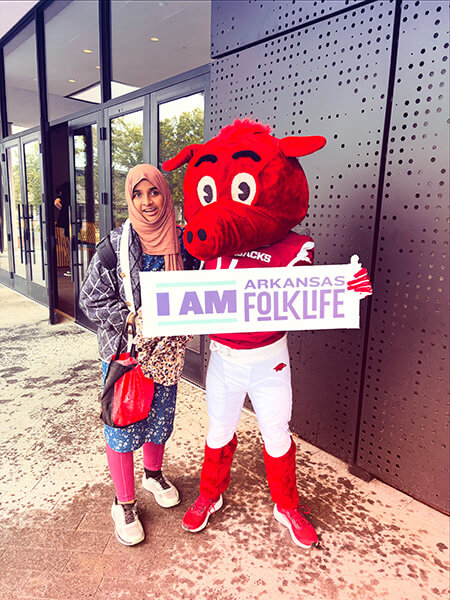 Person wearing a pink hijab and patterned clothes standing next to a red razorback mascot holding a sign that says 'I AM ARKANSAS FOLKLIFE'.