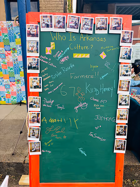Green chalkboard with orange frame titled 'Who Is Arkansas Culture?' surrounded by Polaroid photos, containing colorful handwritten names and words related to Arkansas culture and people.