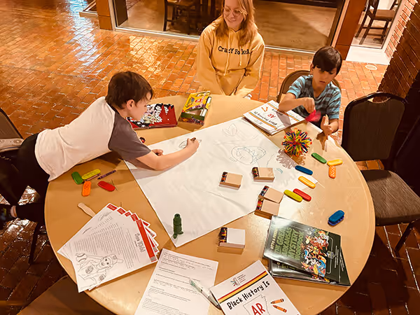 Two boys coloring on a large paper at a round table filled with crayons, books, and activity sheets while a woman watches.