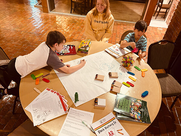 Two boys coloring on a large paper at a round table filled with crayons, books, and activity sheets while a woman watches.