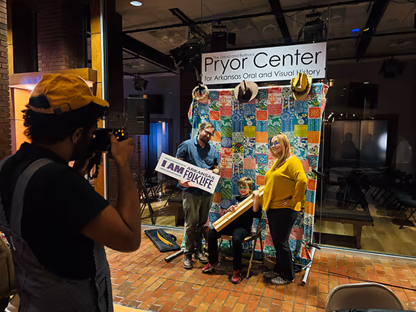 Photographer taking a picture of three people posing at the Pryor Center for Arkansas Oral and Visual History with a colorful quilt backdrop.