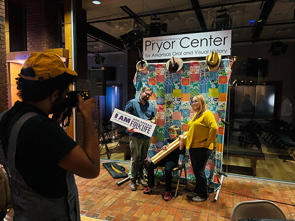 Photographer taking a picture of three people posing at the Pryor Center for Arkansas Oral and Visual History with a colorful quilt backdrop.
