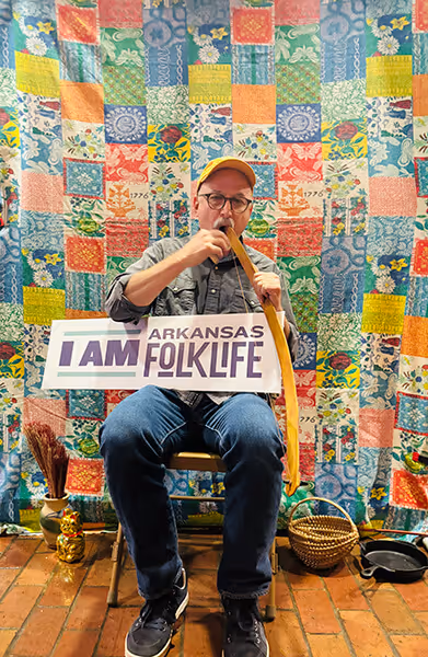 Man wearing a cap seated on a chair playing a mouthbow musical instrument, holding a sign that says 'I AM Arkansas Folk Life' in front of a colorful patchwork quilt backdrop.