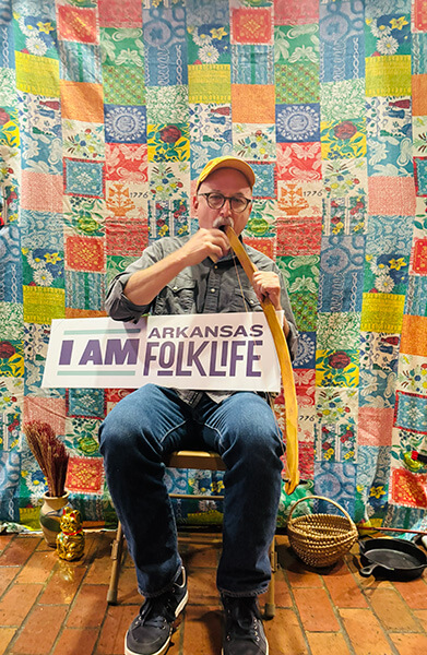 Man wearing a cap seated on a chair playing a mouthbow musical instrument, holding a sign that says 'I AM Arkansas Folk Life' in front of a colorful patchwork quilt backdrop.