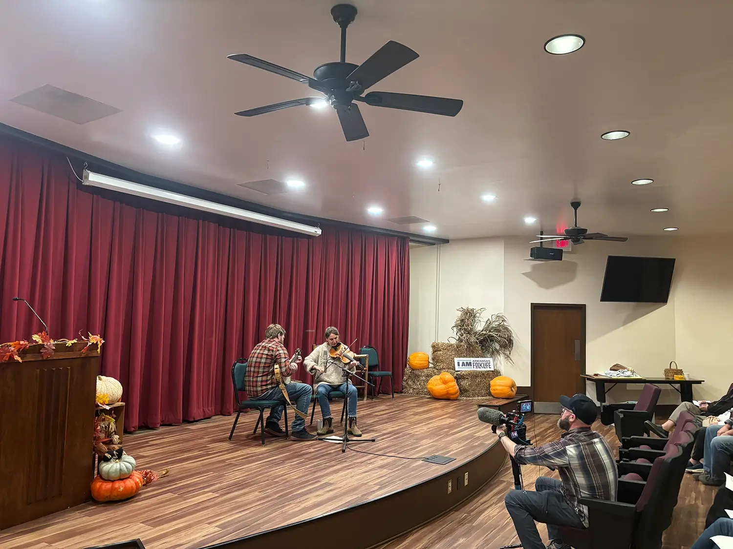 Two musicians playing string instruments on a stage decorated with pumpkins and autumn leaves, while a cameraman films from the audience.