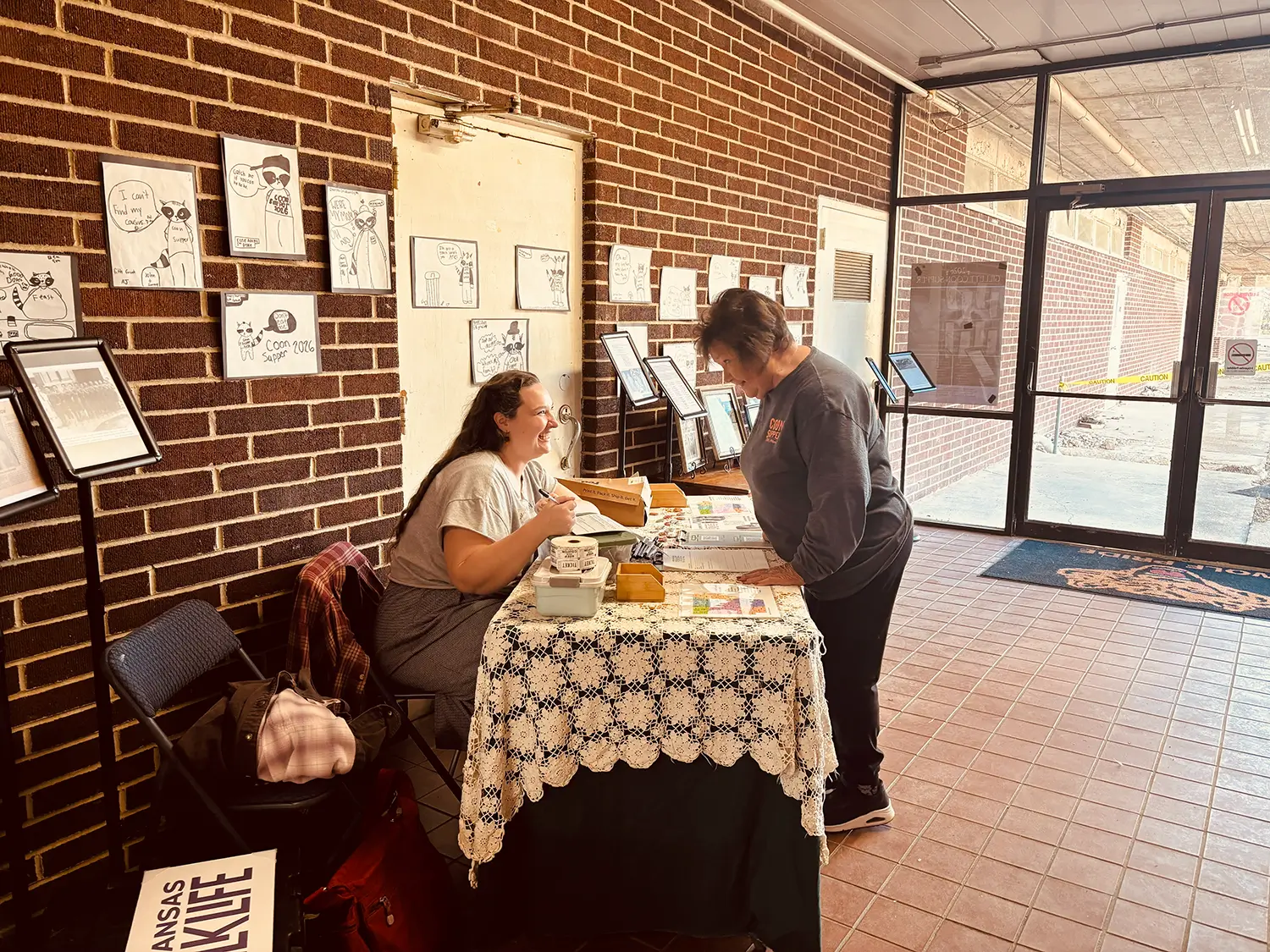 Two women engaged in conversation at a table covered with a lace tablecloth, displaying art and informational materials against a brick wall in a well-lit entryway.