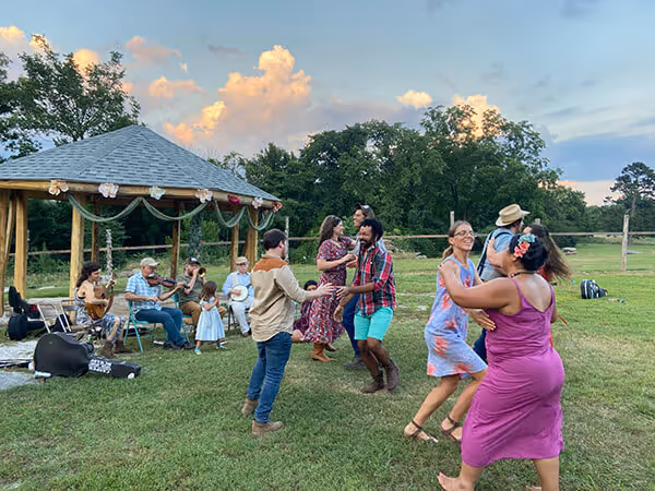 Group of people dancing outdoors near a gazebo with musicians playing instruments in the background during sunset.