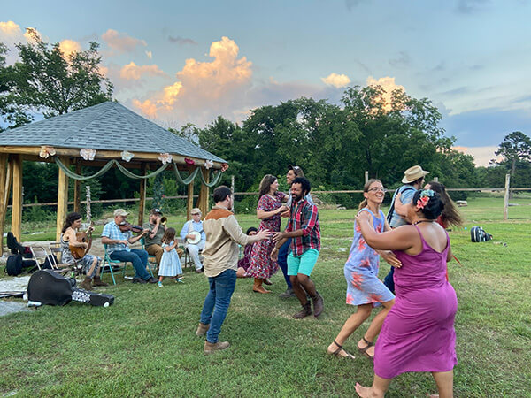 Group of people dancing outdoors near a gazebo with musicians playing instruments in the background during sunset.