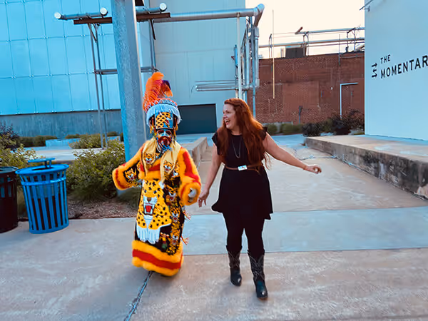 A person in a colorful, detailed traditional costume walking and holding hands with a smiling woman in black dress and boots outdoors.