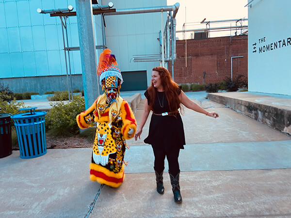 A person in a colorful, detailed traditional costume walking and holding hands with a smiling woman in black dress and boots outdoors.