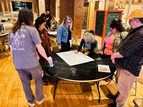 A group of six people standing around a table with a large sheet of paper, engaged in discussion and writing in an indoor setting with brick pillars and wooden floors.