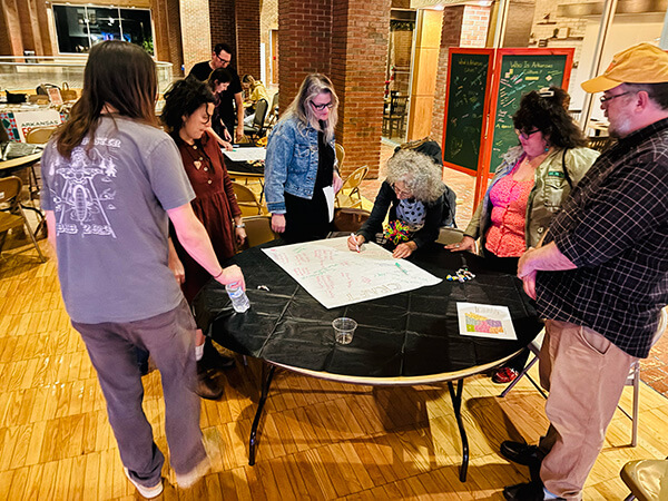 A group of six people standing around a table with a large sheet of paper, engaged in discussion and writing in an indoor setting with brick pillars and wooden floors.