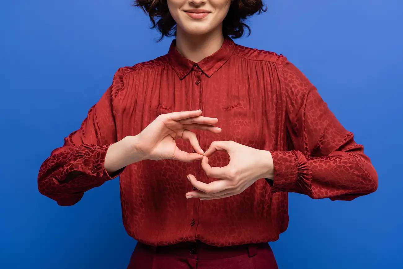 Woman in a red blouse smiling and signing with both hands against a blue background.