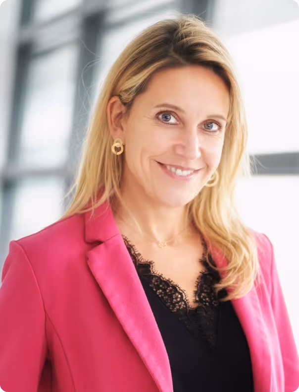 Porträt einer lächelnden Frau mit langen blonden Haaren, die eine pinke Jacke und ein schwarzes Spitzenoberteil trägt.