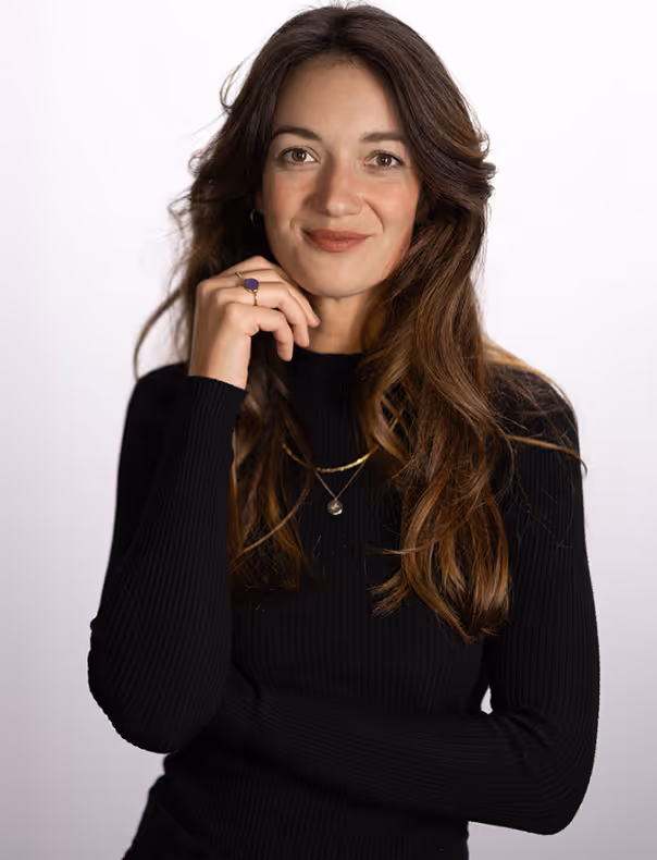 Porträt einer lächelnden Frau mit langen braunen Haaren, schwarzen Pullover und goldenen Halsketten vor weißem Hintergrund.