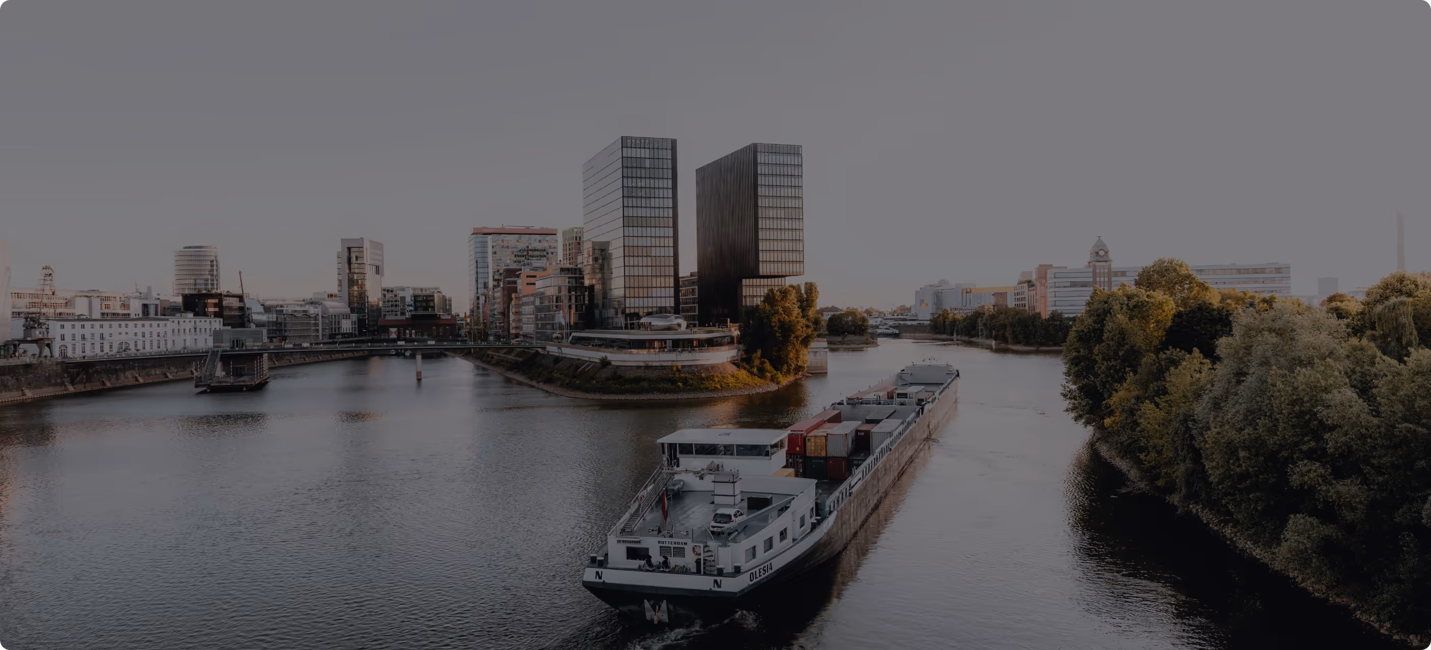 Frachtschiff auf einem Fluss, umgeben von modernen Gebäuden und Bäumen bei Sonnenuntergang.