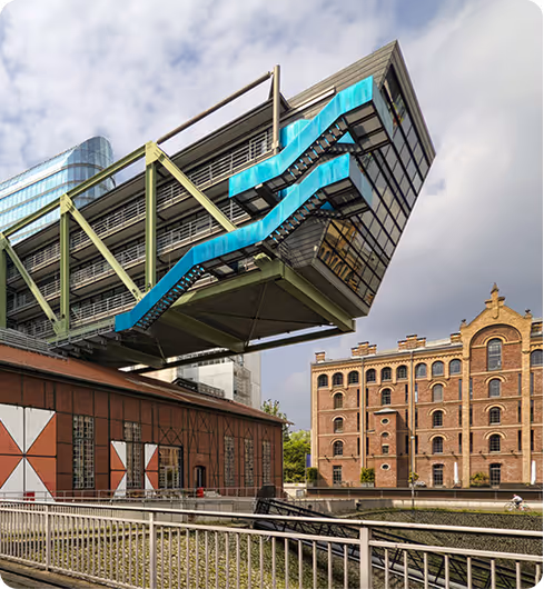 Modernes Gebäude mit auffälliger blauer, gezackter Außentreppe über einem Backsteingebäude neben einem Kanal in der Stadt.