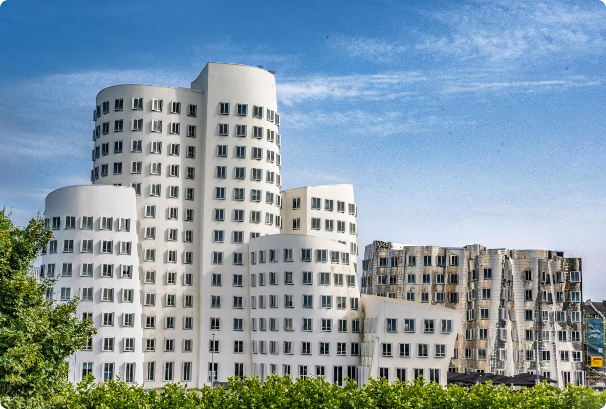 Mehrere moderne, weiße Gebäude mit gebogenen Formen und vielen Fenstern vor blauem Himmel und grünen Bäumen.