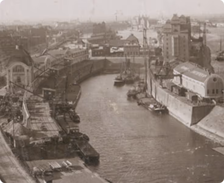 Historisches Schwarz-Weiß-Foto eines belebten Hafens mit Lagerhäusern, Kränen und Schiffen am Flussufer.