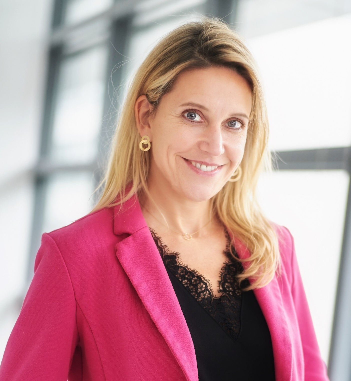 Lächelnde blonde Frau mit pinkem Blazer und schwarzer Spitzenbluse vor hellem Fenster.