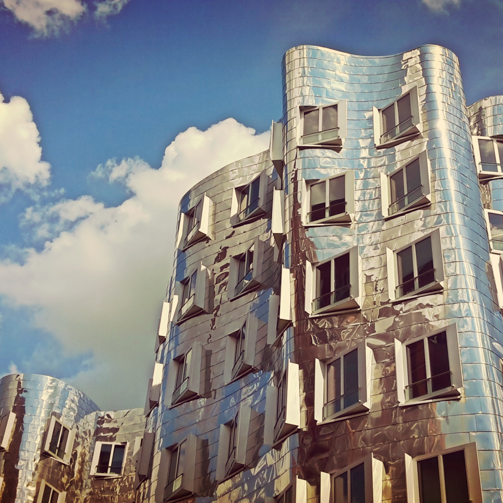 Gebäude mit wellenförmiger, reflektierender Metallfassade und vielen Fenstern vor blauem Himmel mit Wolken.