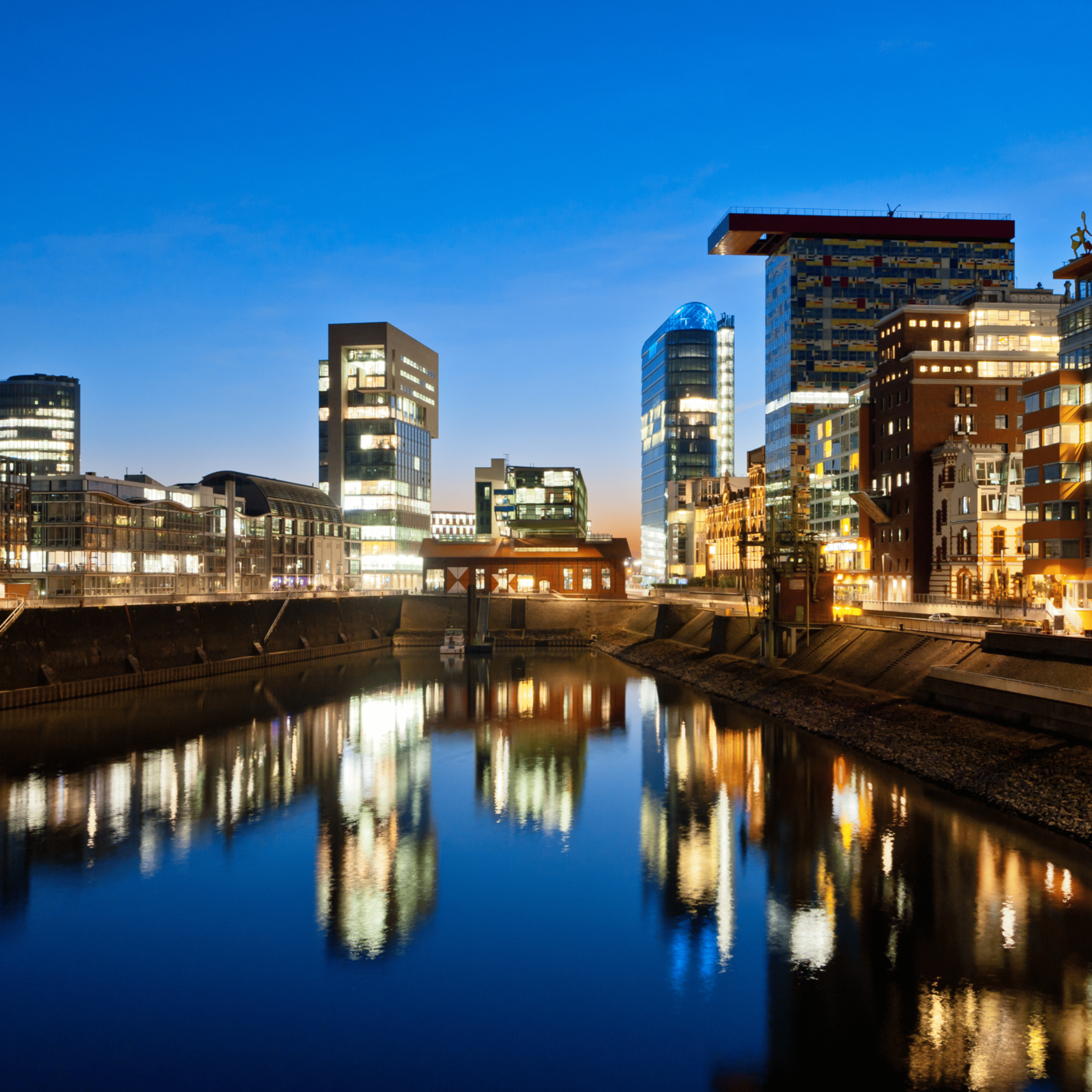 Nächtliche Skyline moderner Gebäude mit Reflexionen im ruhigen Wasser eines Hafens.