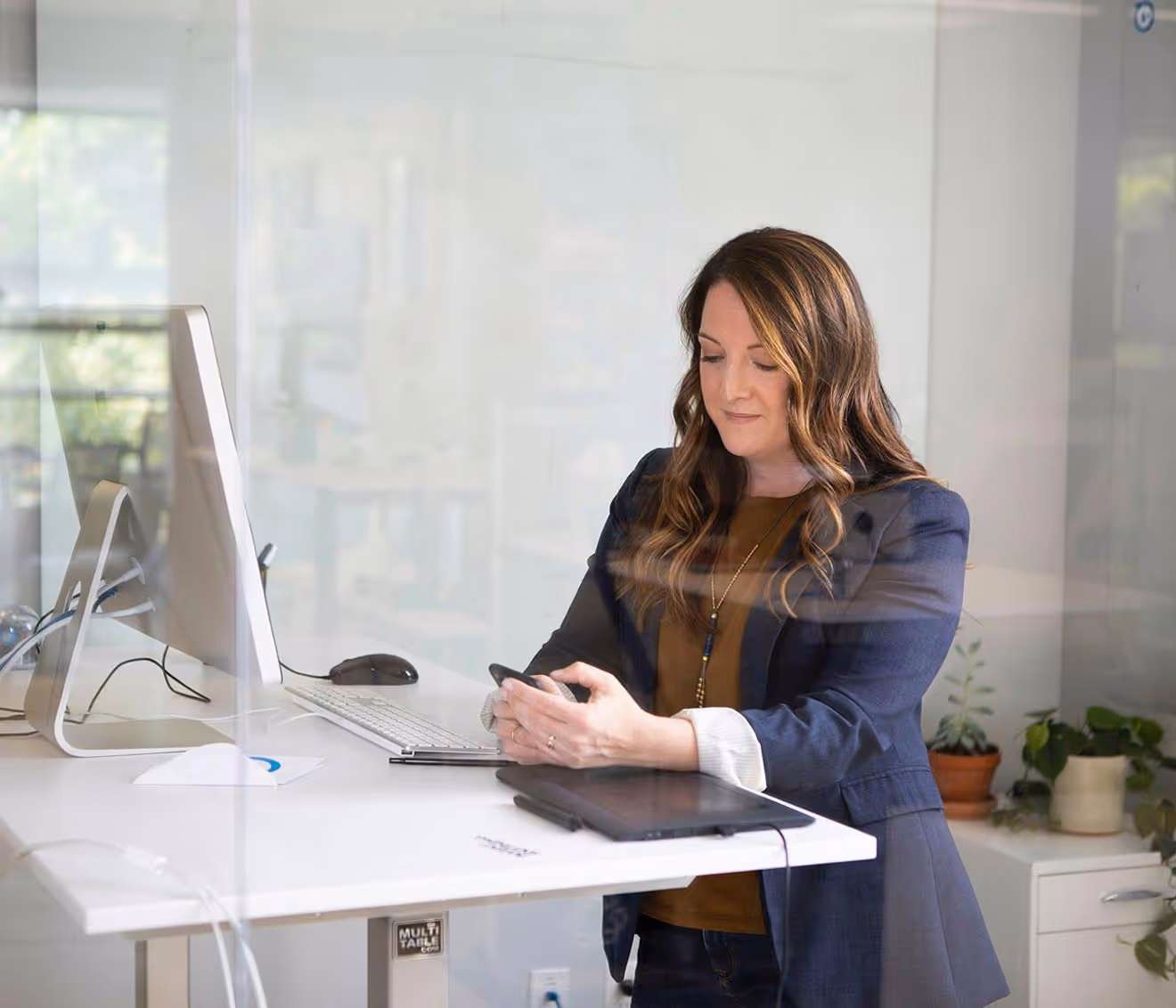 Woman in a navy blazer using a smartphone while standing at a white desk with a computer and plants in the background.