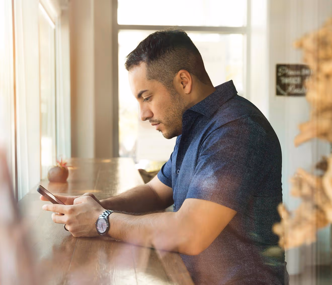 Man sitting at a wooden table by a window, looking intently at his smartphone.