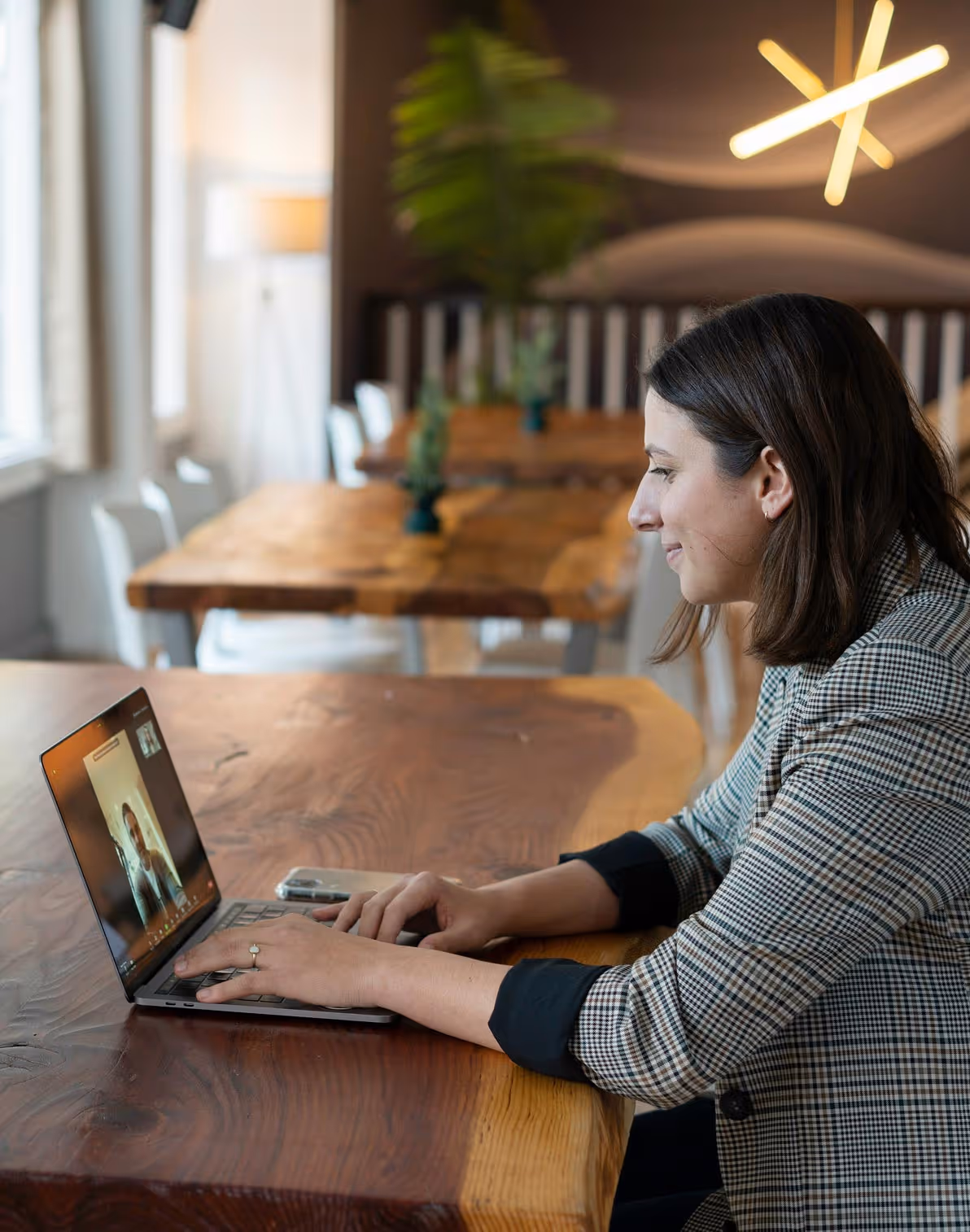 Woman in a checkered blazer engaged in a video call on a laptop at a wooden table in a modern office setting.