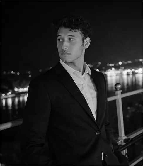 Black and white photo of a young man in a suit looking to his left with city lights in the background.