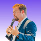 Man with curly hair and beard speaking into a microphone against a purple background.