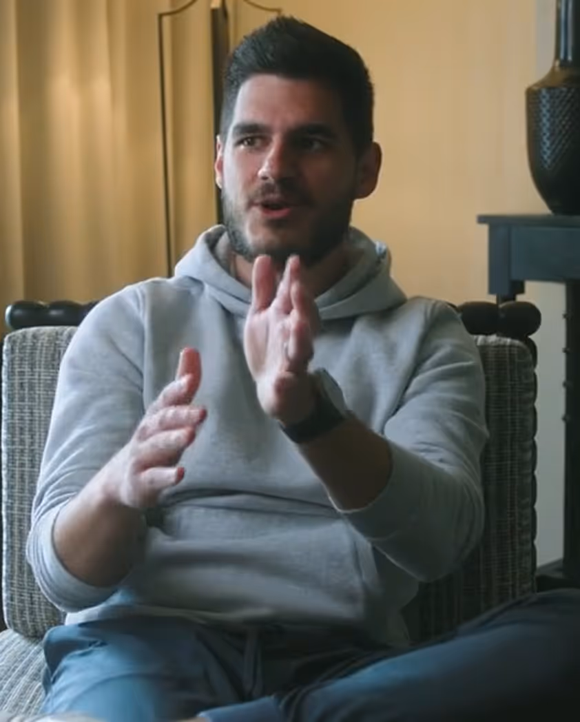 Bearded man in a gray hoodie gesturing with hands while sitting on a chair indoors.