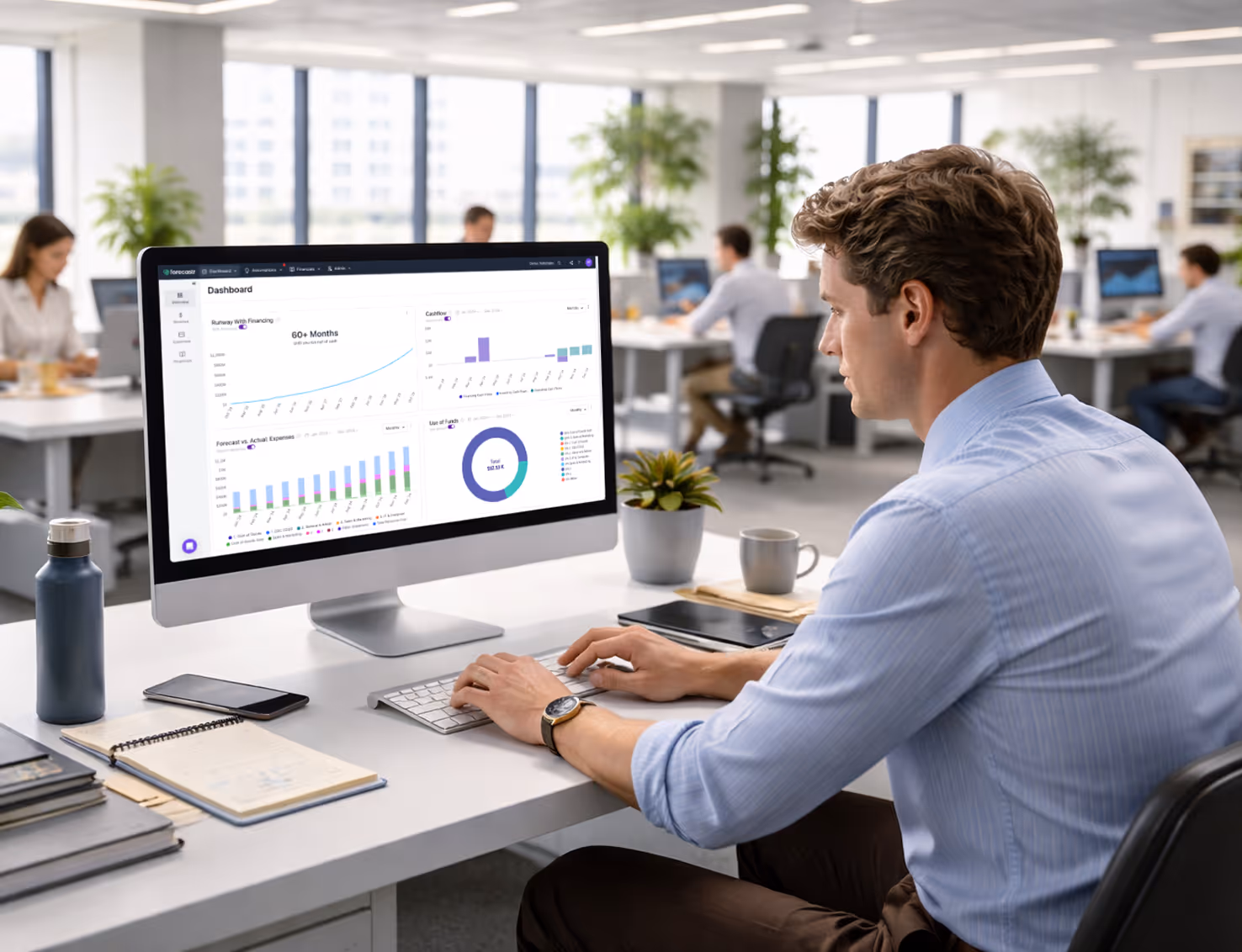 Man in blue shirt working on desktop with dashboard charts in a modern open office space.