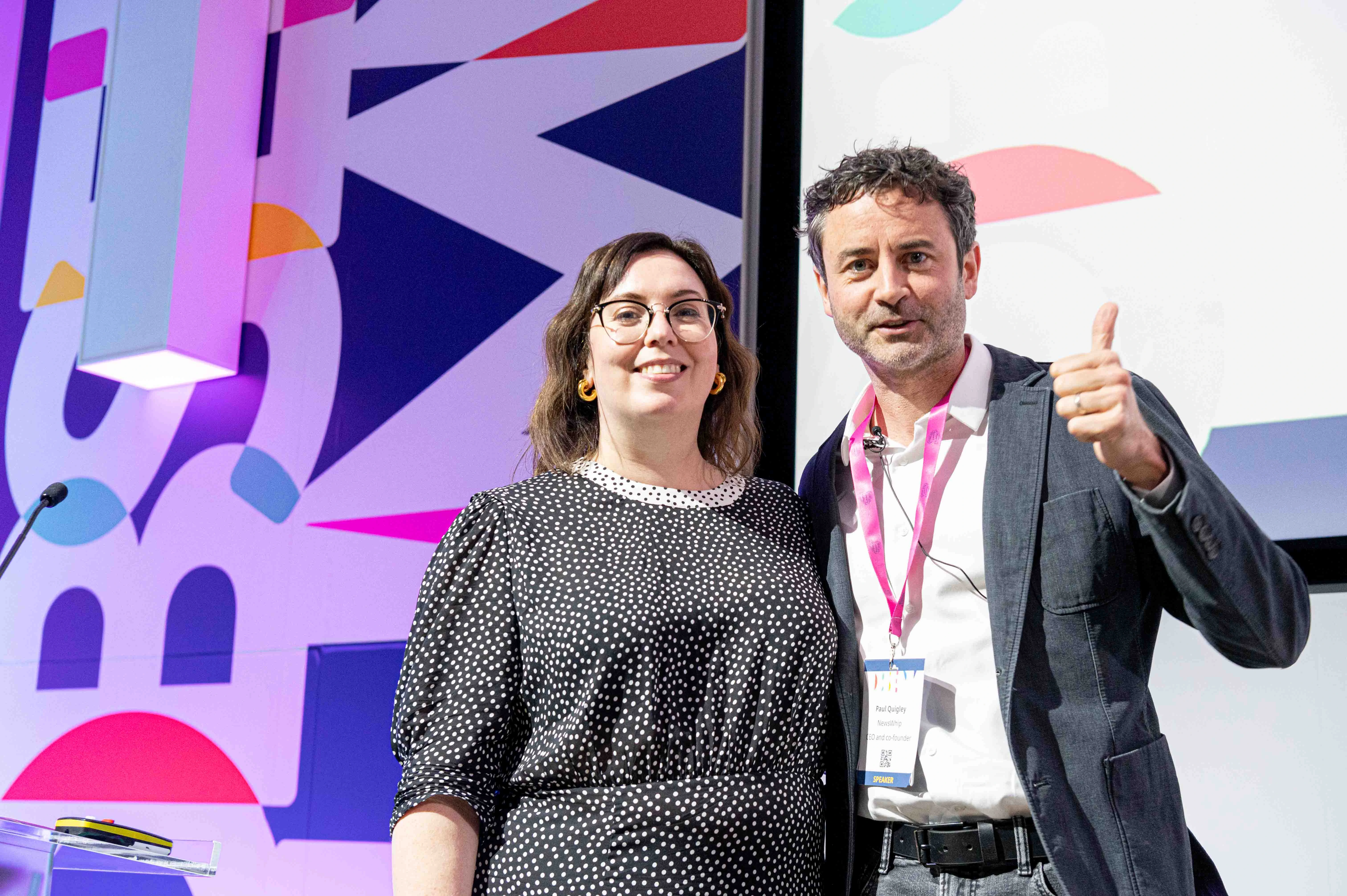 Woman and man smiling at event, man giving thumbs up with speaker badge on lanyard.