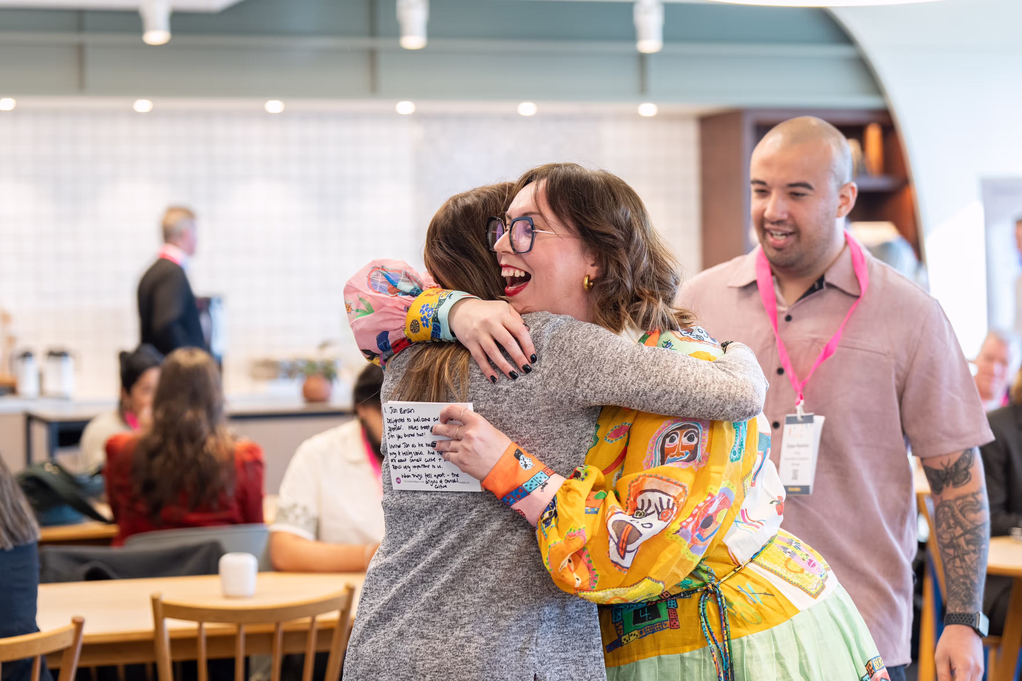 Two women warmly hugging in a lively indoor setting while a man with tattoos looks on smiling.
