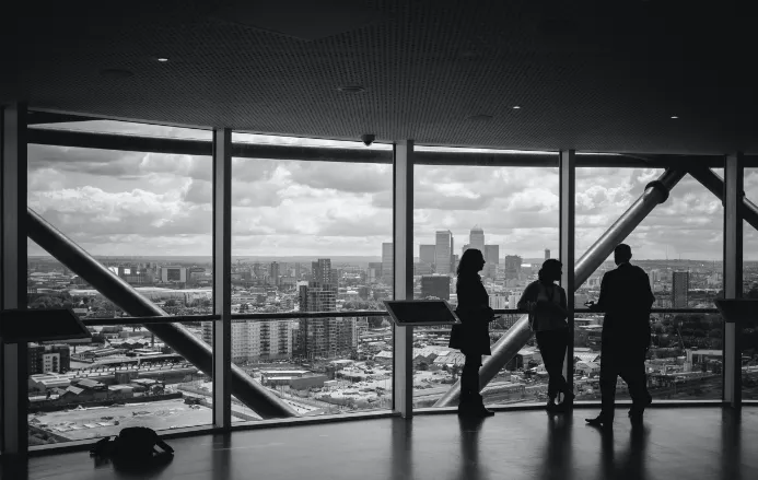 Silhouettes of three people standing inside a high-rise building overlooking a cityscape through large windows.