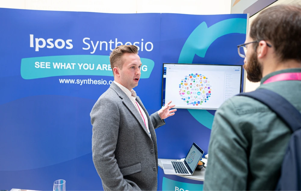 Man in gray blazer presenting data visualizations on screens at Ipsos Synthesio booth.