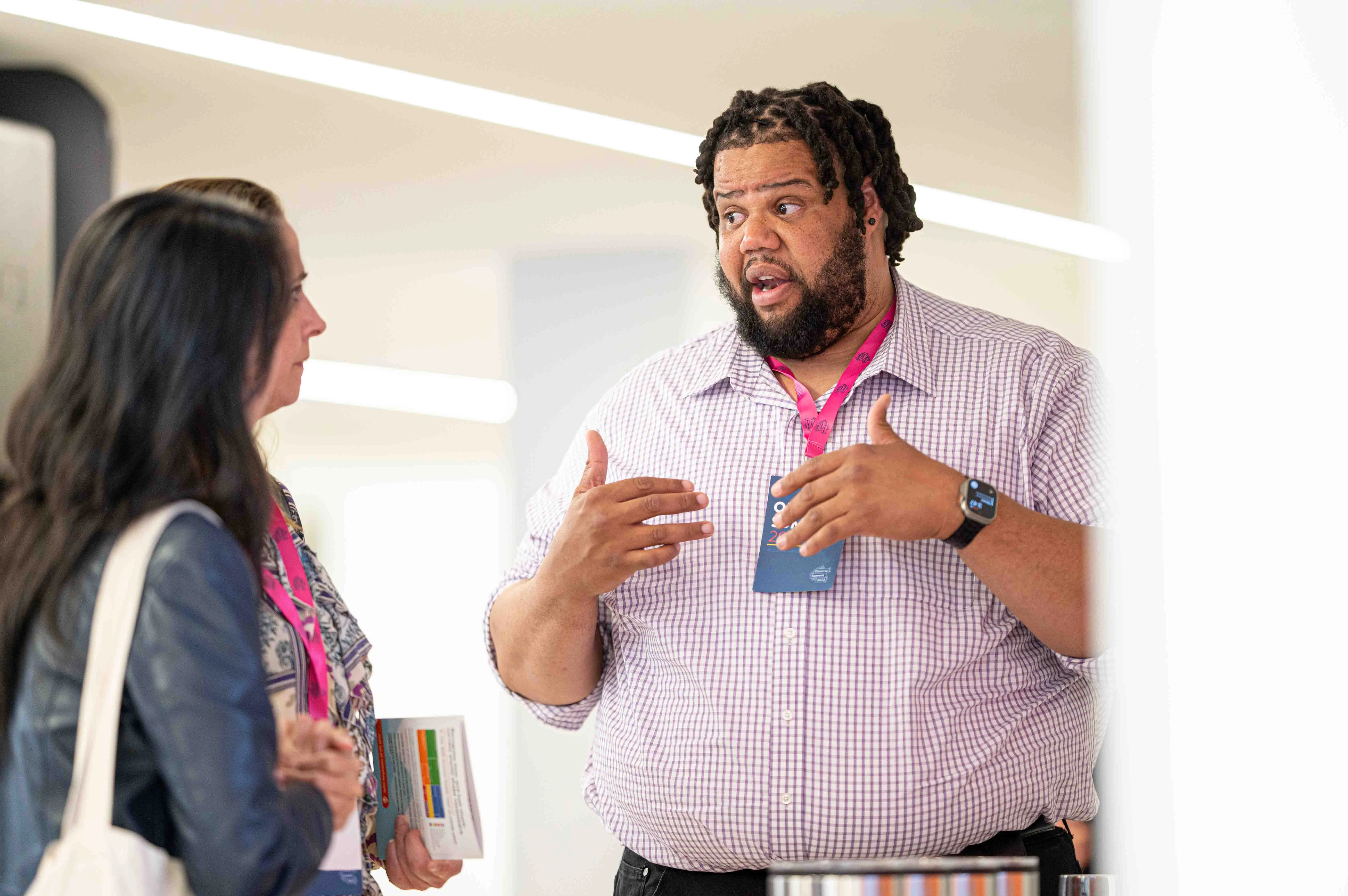 Man with lanyard speaking animatedly to two women holding papers in a bright indoor setting.