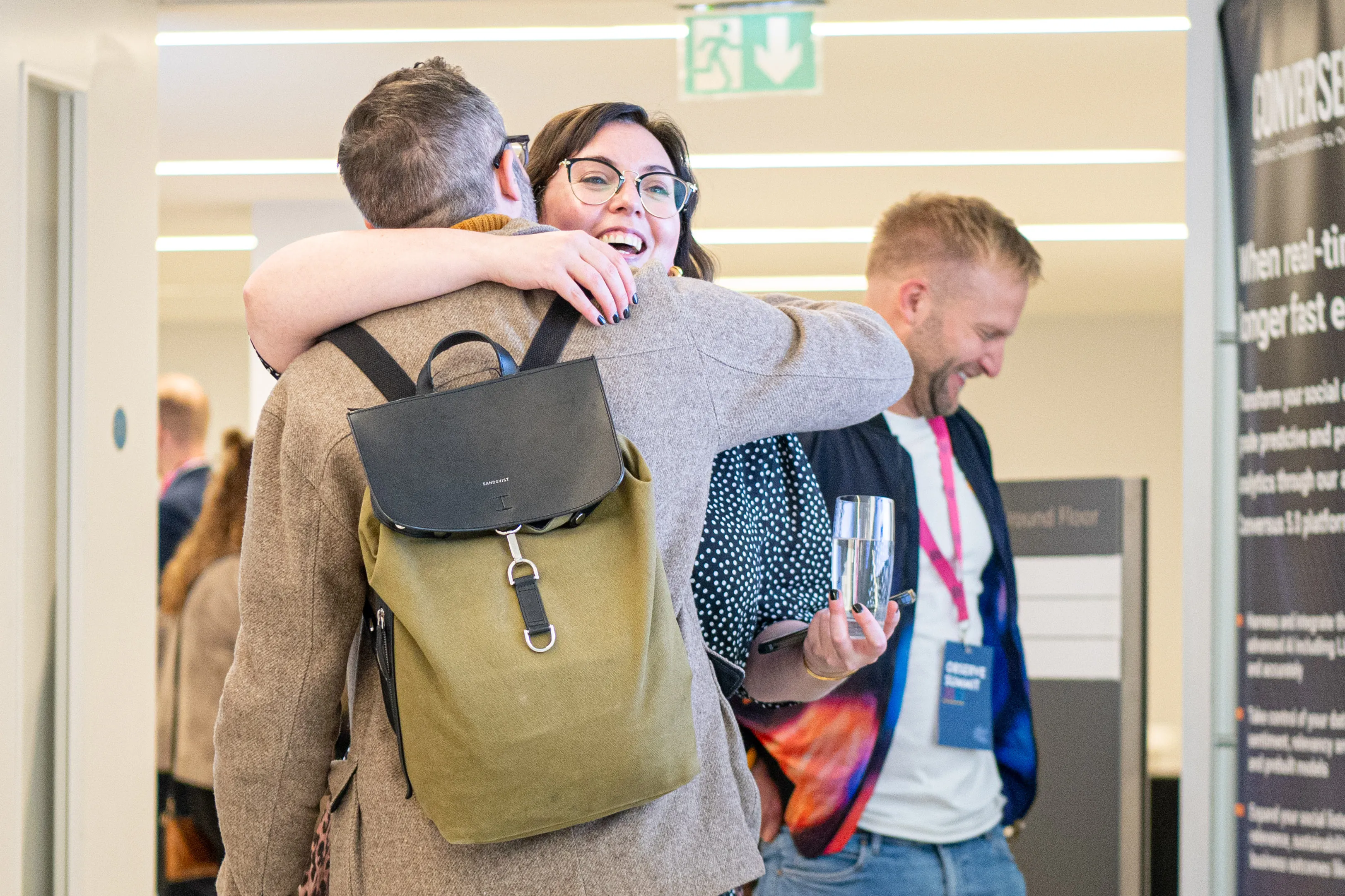 Two people warmly hugging in a bright indoor space with another person smiling nearby.
