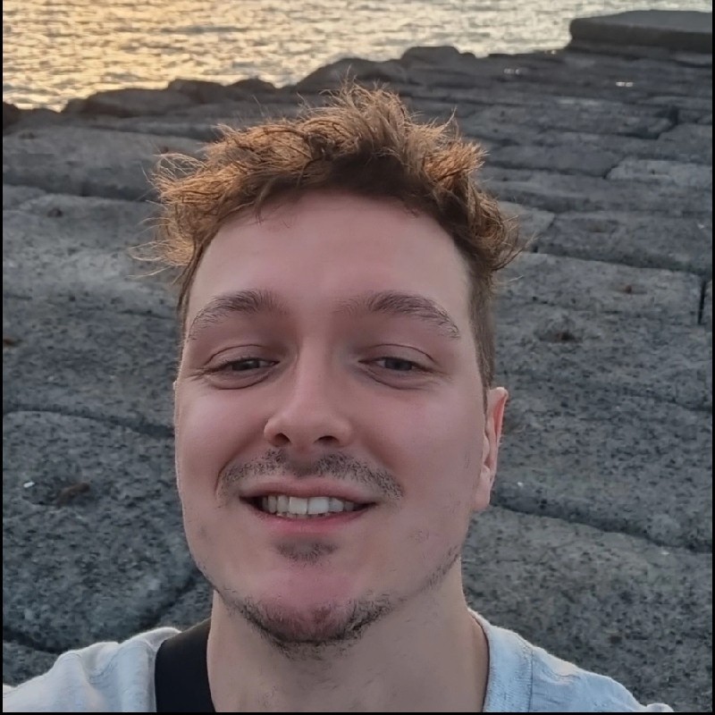 Smiling young man with curly hair taking a selfie on rocky ground near water at sunset.