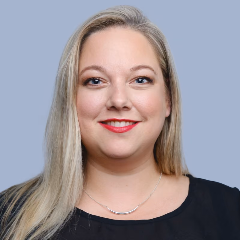 Smiling blonde woman with red lipstick wearing a black top and a silver necklace against a blue background.
