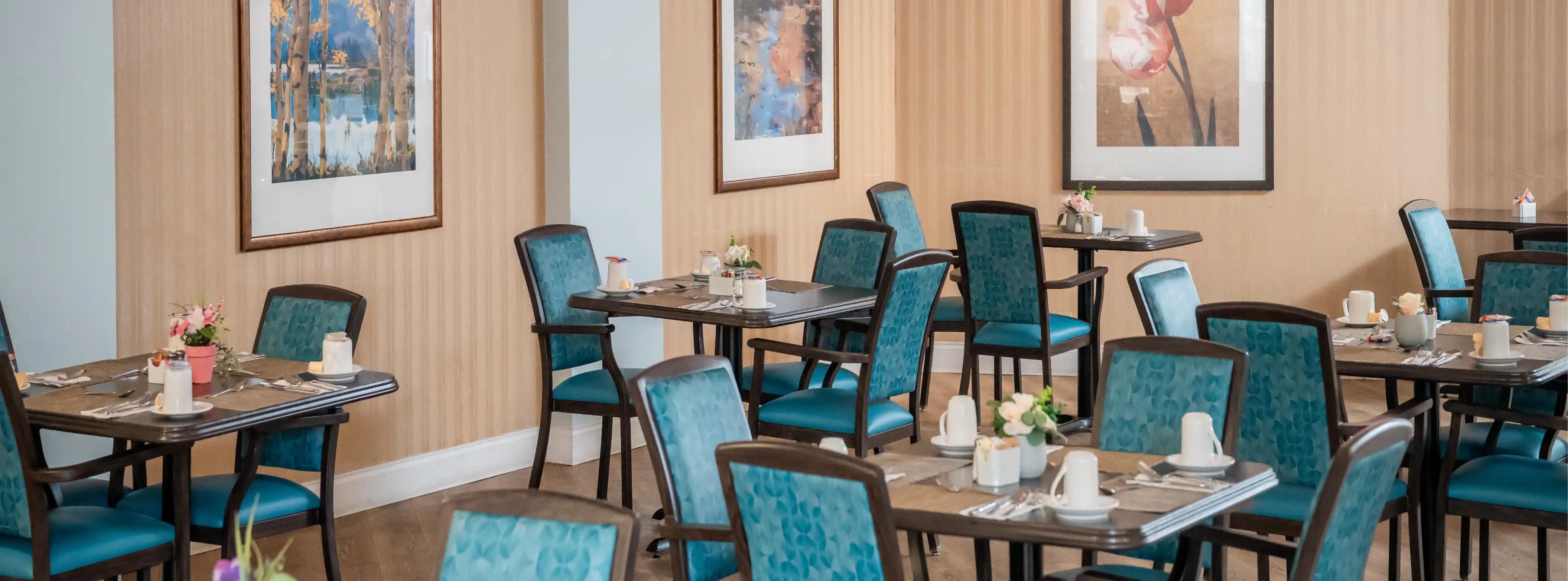 Dining area with wooden tables set with white cups, silverware, and small flower arrangements, surrounded by teal upholstered chairs.