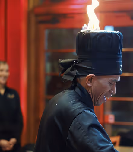 Man wearing a black traditional outfit and tall black hat with a flame on top, smiling in an indoor setting with red walls and blurred figure in the background.